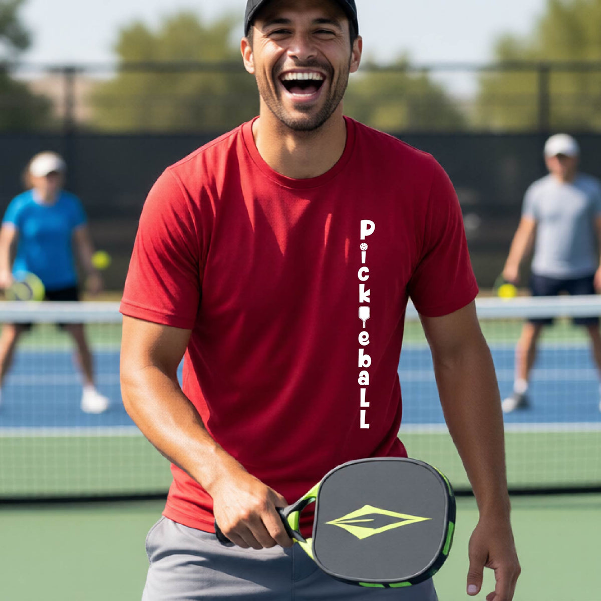 A man wearing a red short-sleeve t-shirt with a white graphic text 'PICKLEBALL' on the front with a pickleball paddle replacing the L, standing on a pickleball court.