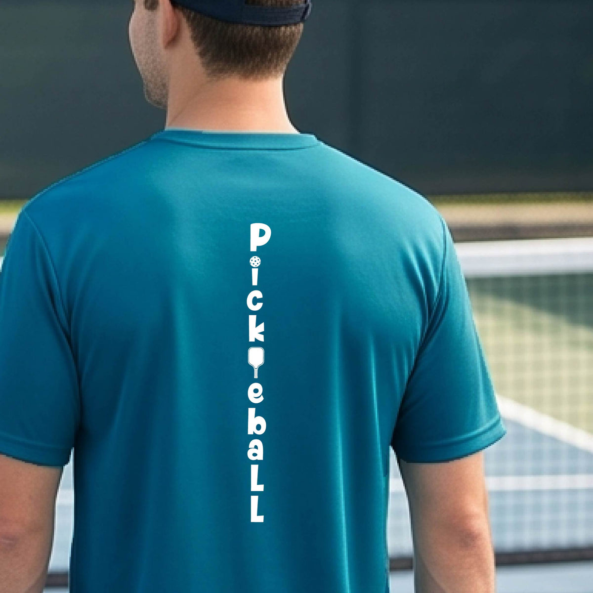 A man wearing a teal short-sleeve t-shirt with a white graphic text 'PICKLEBALL' on the back with a pickleball paddle replacing the L, standing on a pickleball court.