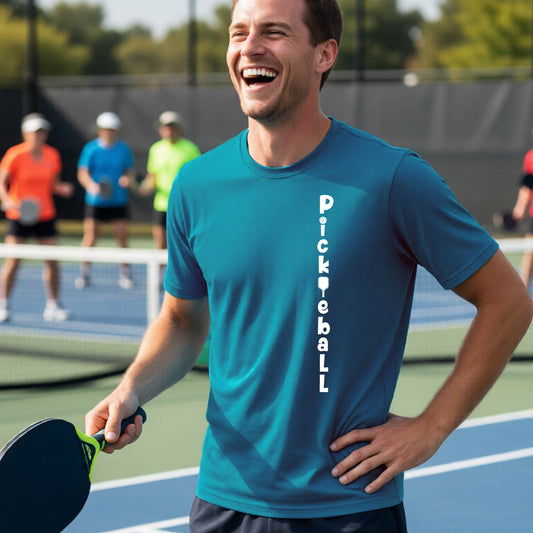 A man wearing a teal short-sleeve t-shirt with a white graphic text 'PICKLEBALL' on the front with a pickleball paddle replacing the L, standing on a pickleball court.