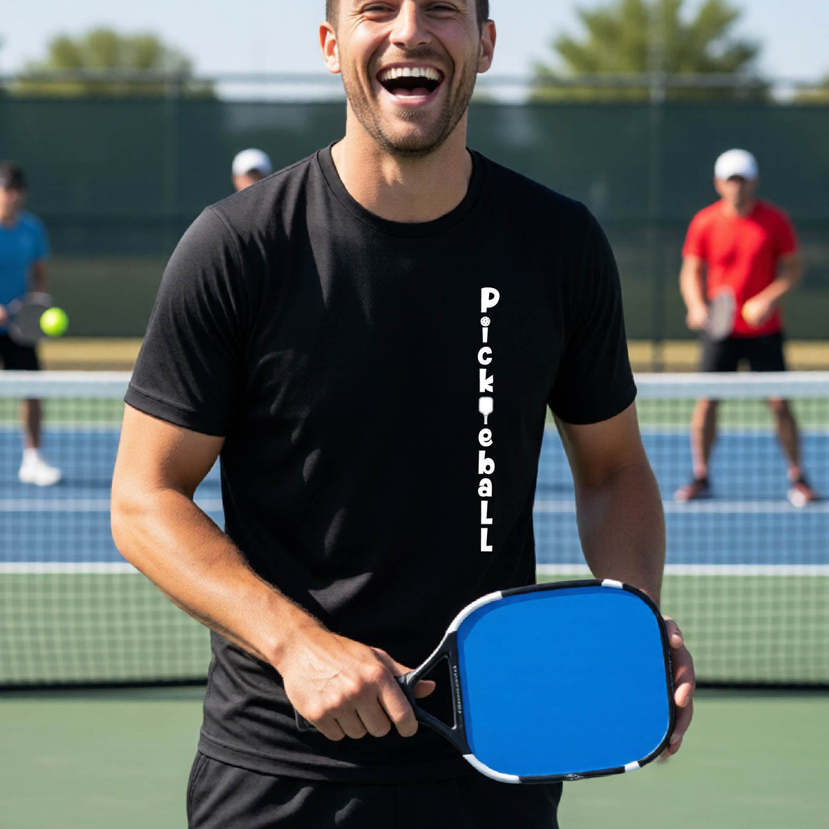 A man wearing a black short-sleeve t-shirt with a white graphic text 'PICKLEBALL' on the front with a pickleball paddle replacing the L, standing on a pickleball court.