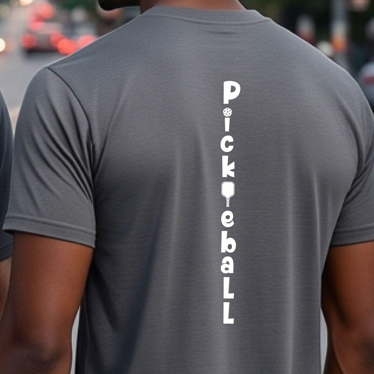 A man wearing a gray short-sleeve t-shirt with a white graphic text 'PICKLEBALL' on the back with a pickleball paddle replacing the L, standing on a pickleball court.