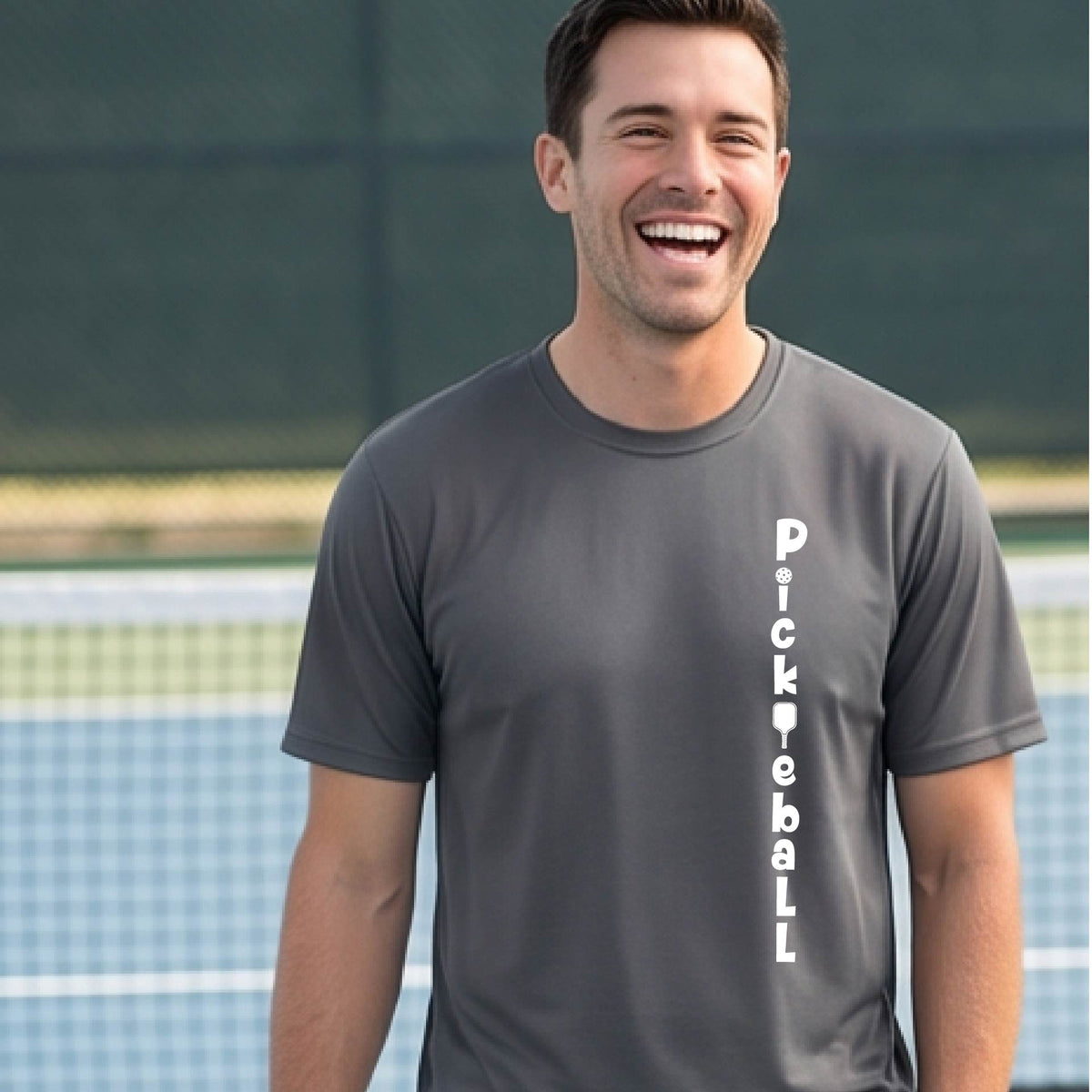 A man wearing a gray short-sleeve t-shirt with a white graphic text 'PICKLEBALL' on the front with a pickleball paddle replacing the L, standing on a pickleball court.