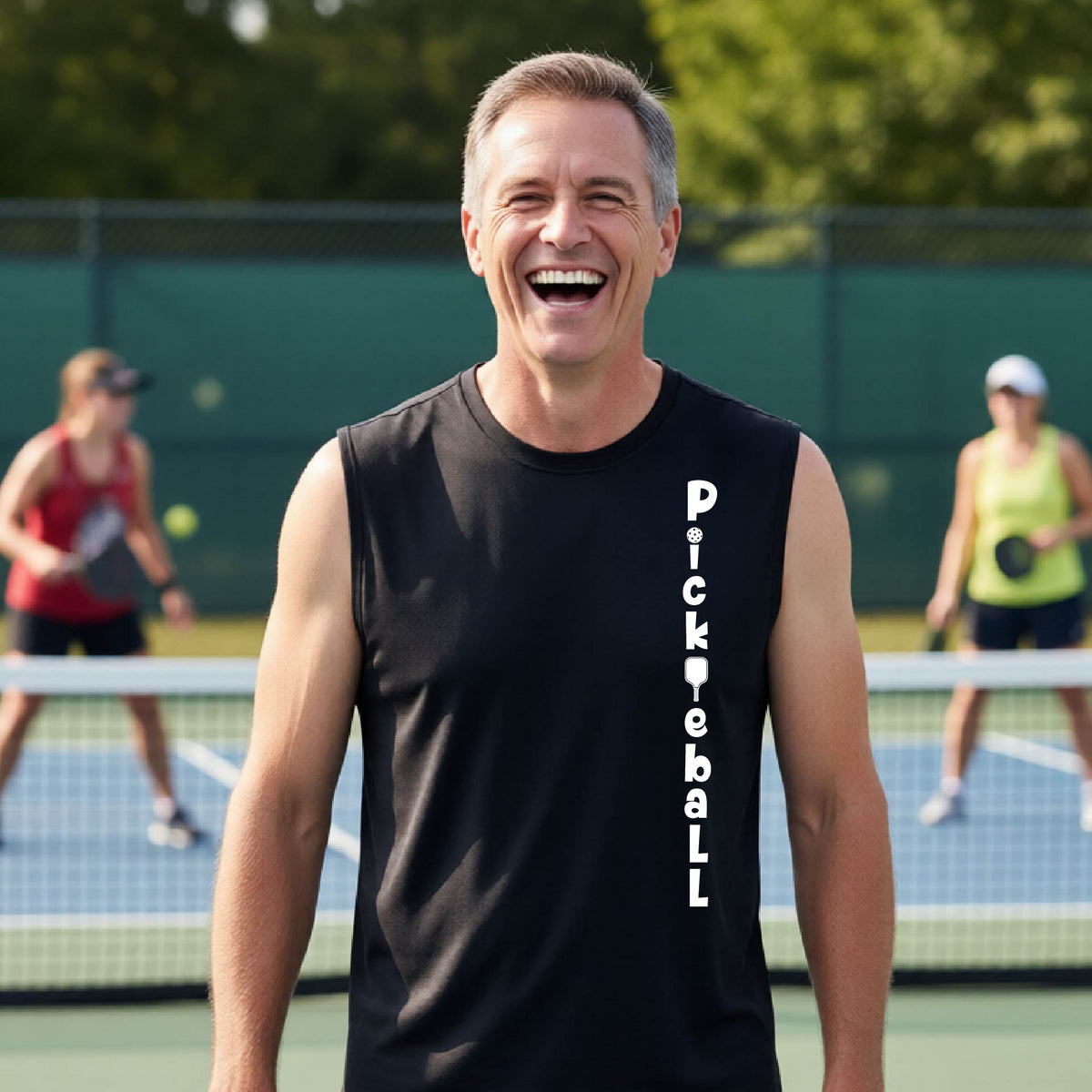 A man wearing a black sleeveless t-shirt with a white graphic text 'PICKLEBALL' on the front with a pickleball paddle replacing the L, standing on a pickleball court
