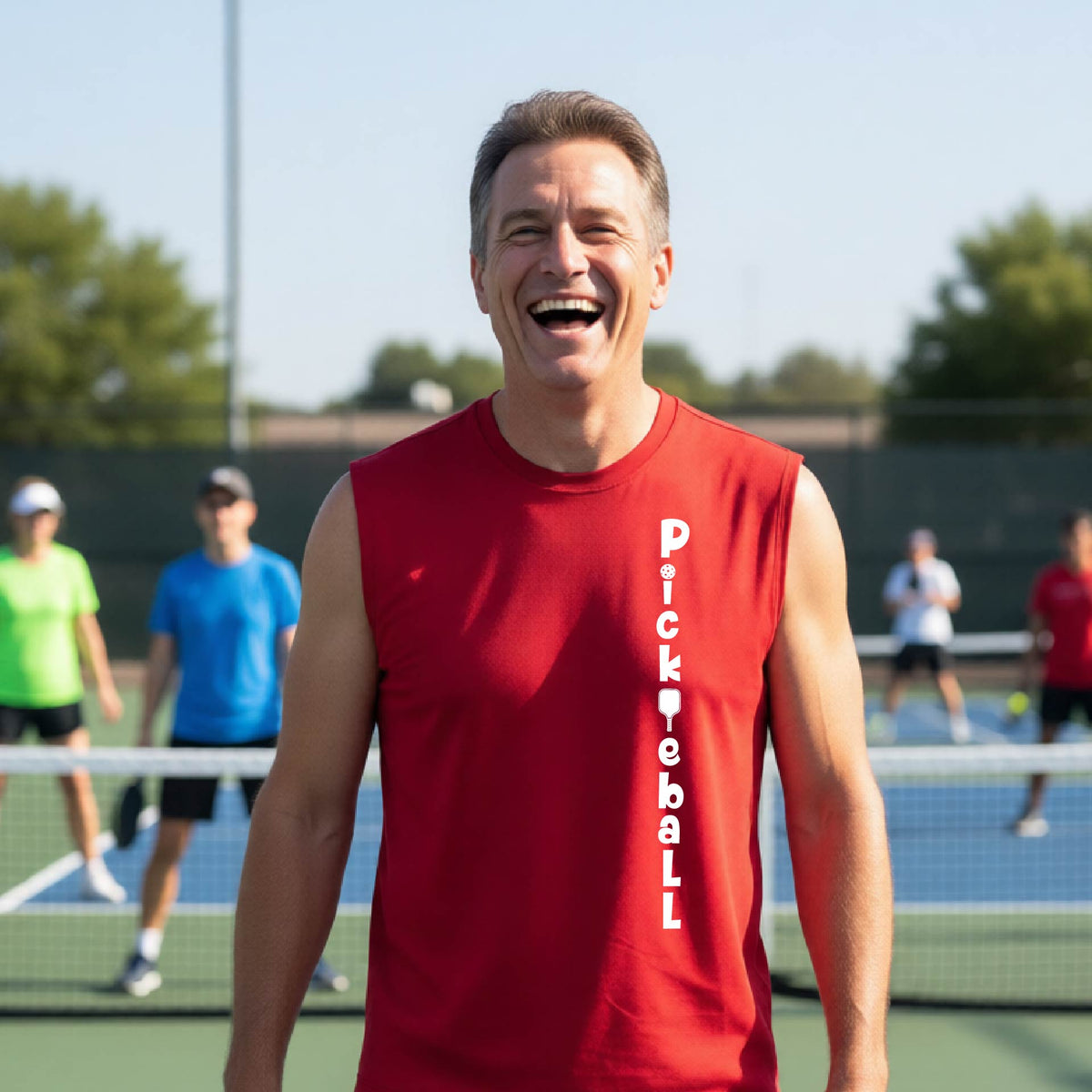 A man wearing a red sleeveless t-shirt with a white graphic text 'PICKLEBALL' on the front with a pickleball paddle replacing the L, standing on a pickleball court