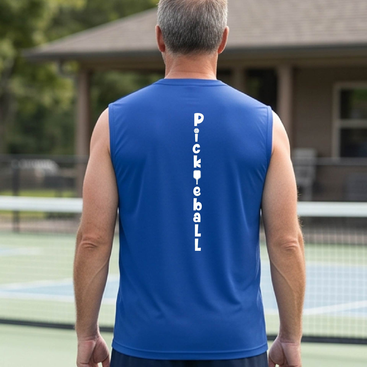 A man wearing a royal blue sleeveless t-shirt with a white graphic text 'PICKLEBALL' on the back with a pickleball paddle replacing the L, standing on a pickleball court