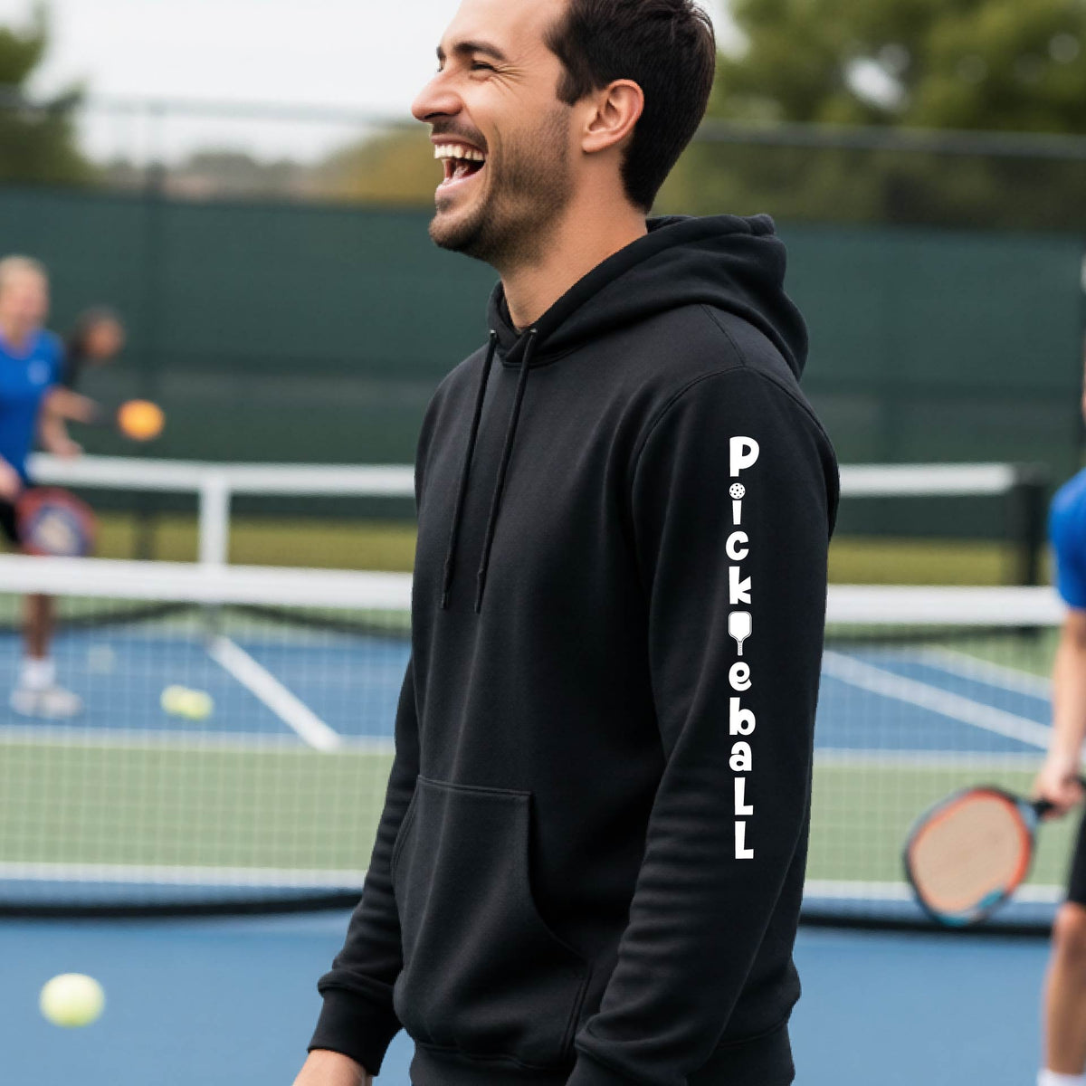 A man wearing a black long-sleeve unisex hoodie pullover with a white graphic text 'PICKLEBALL' on the sleeve with a pickleball paddle replacing the L, standing on a pickleball court.