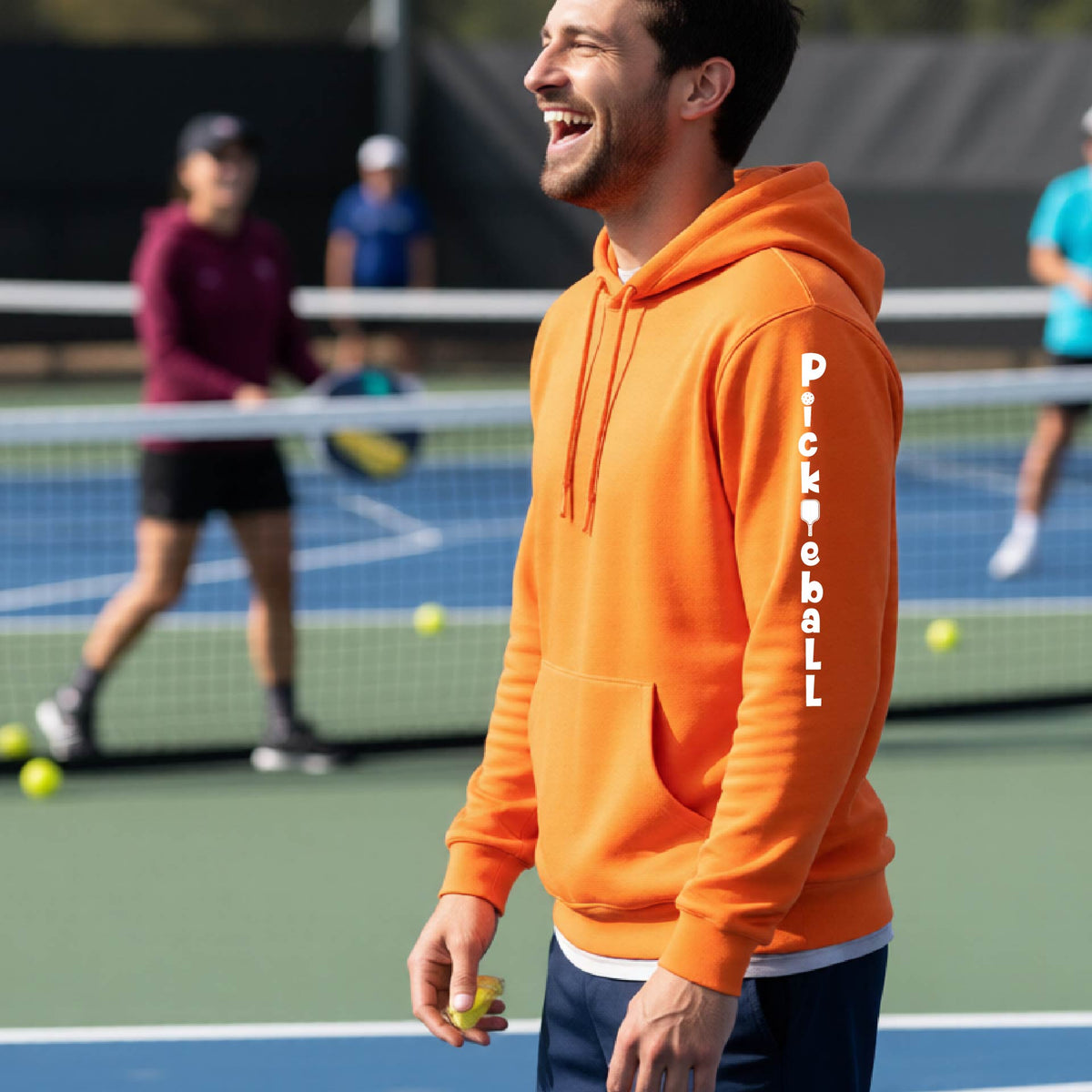 A man wearing an orange long-sleeve unisex hoodie pullover with a white graphic text 'PICKLEBALL' on the sleeve with a pickleball paddle replacing the L, standing on a pickleball court.