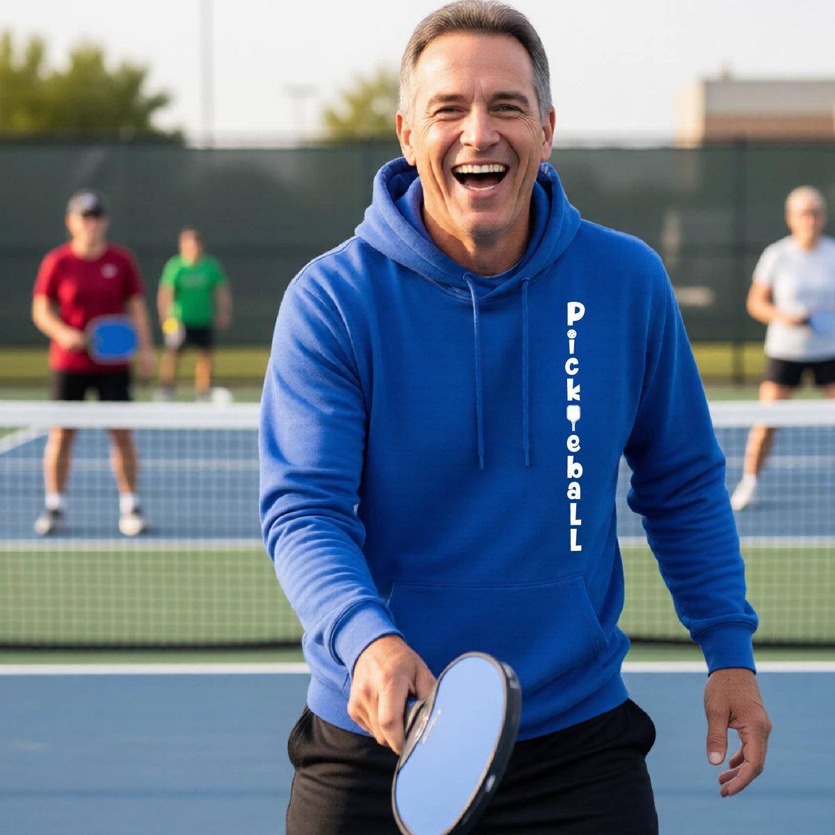 A man wearing a royal blue long-sleeve unisex hoodie pullover with a white graphic text 'PICKLEBALL' on the front with a pickleball paddle replacing the L, standing on a pickleball court.