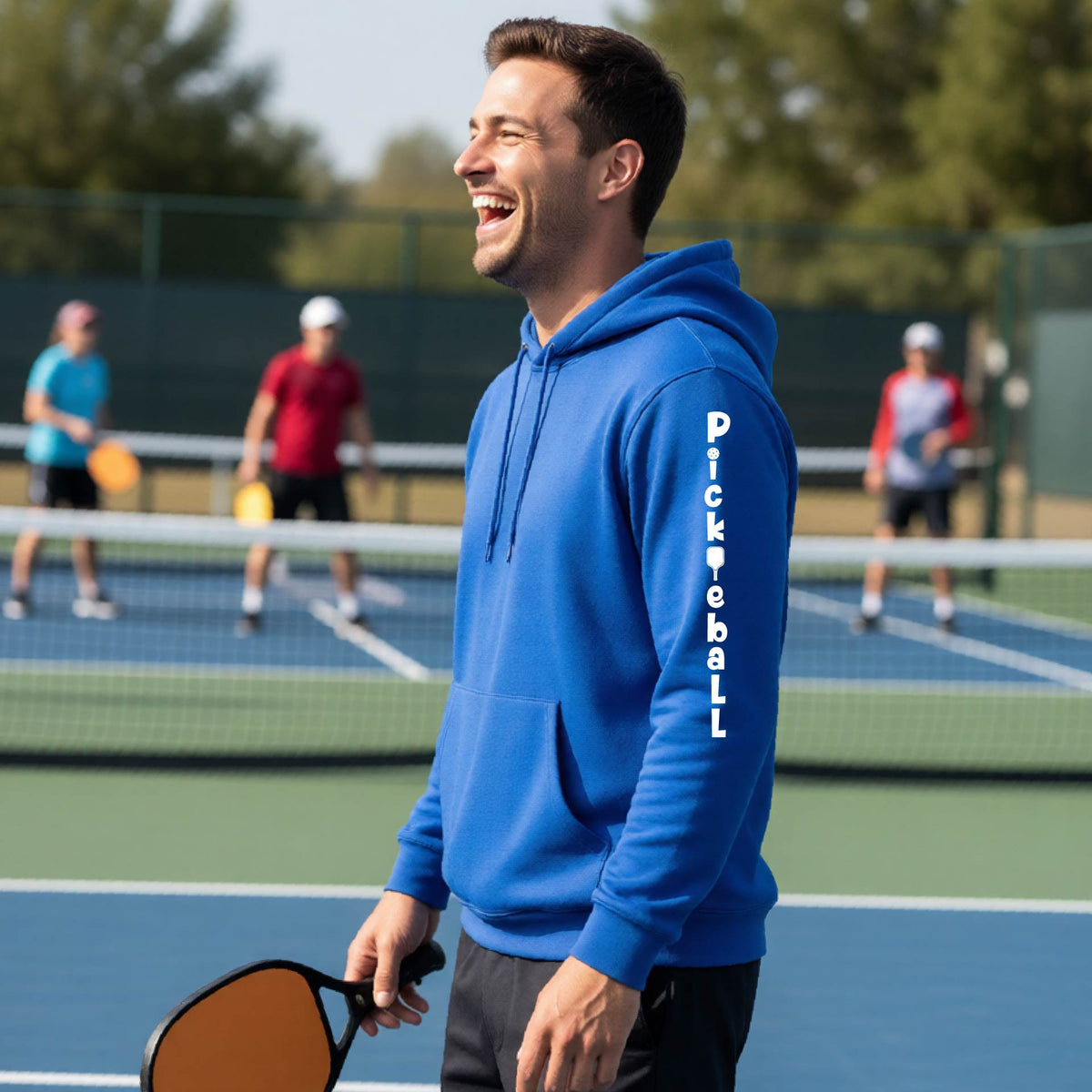 A man wearing a royal blue long-sleeve unisex hoodie pullover with a white graphic text 'PICKLEBALL' on the sleeve with a pickleball paddle replacing the L, standing on a pickleball court.