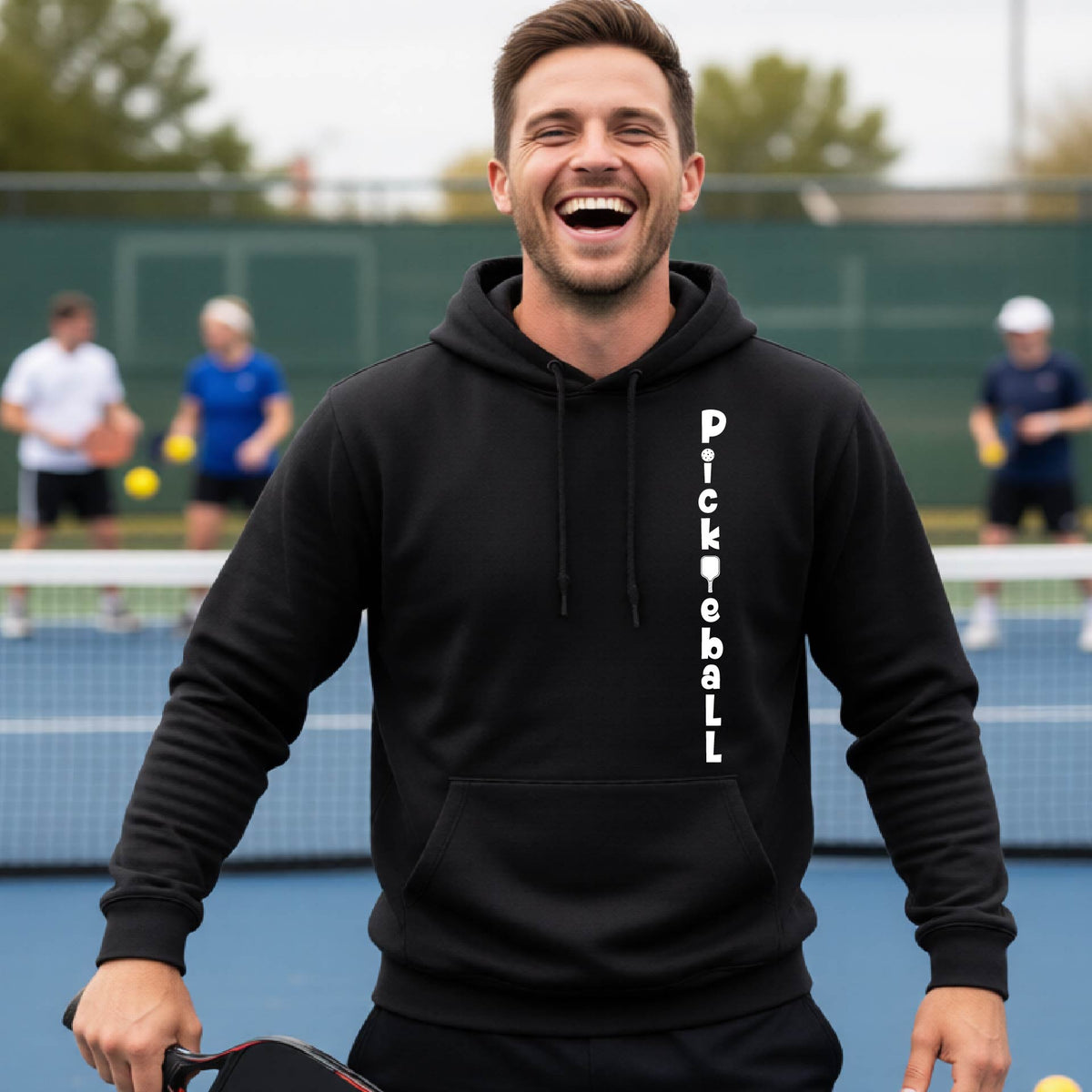 A man wearing a black long-sleeve unisex hoodie pullover with a white graphic text 'PICKLEBALL' on the front with a pickleball paddle replacing the L, standing on a pickleball court.