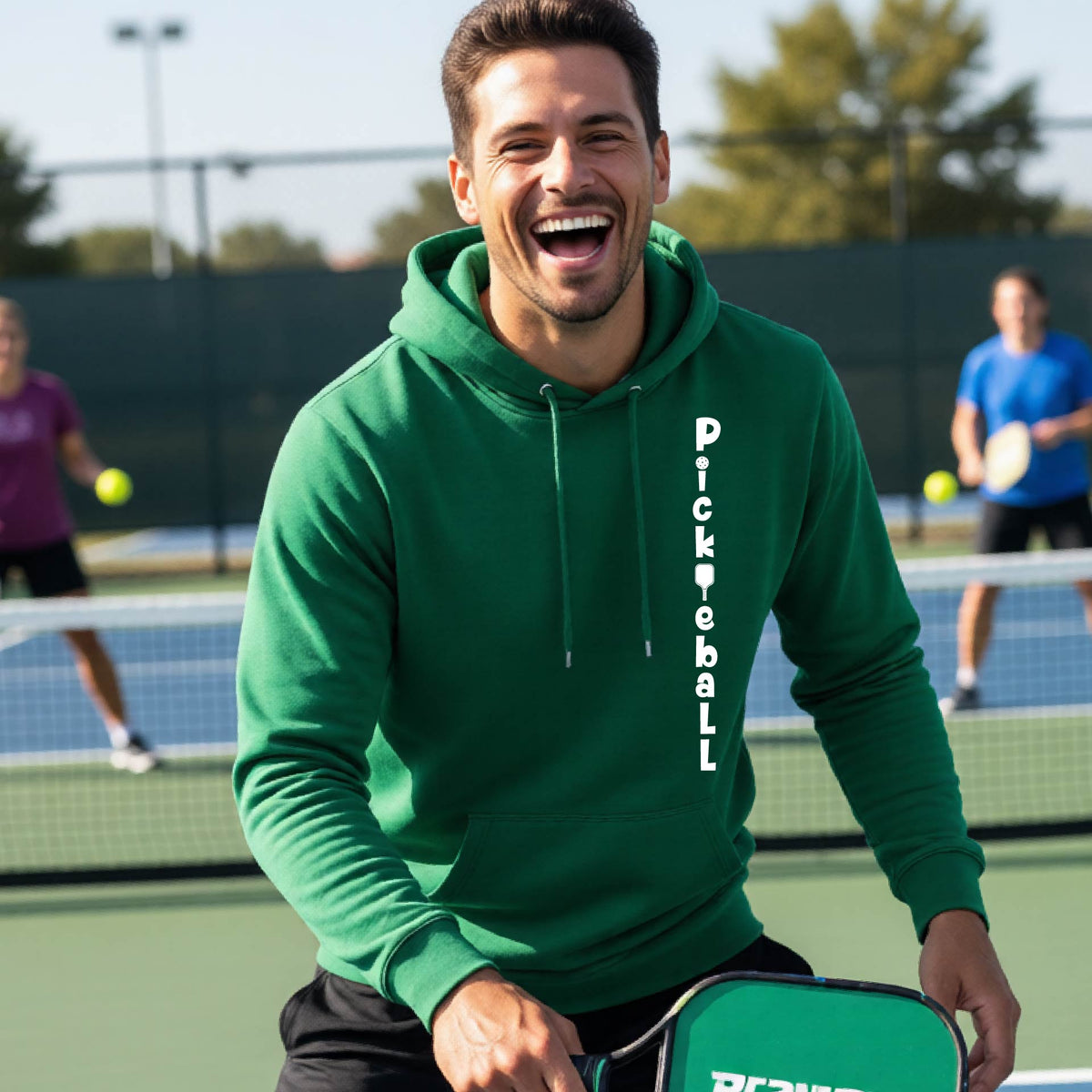 A man wearing a green long-sleeve unisex hoodie pullover with a white graphic text 'PICKLEBALL' on the front with a pickleball paddle replacing the L, standing on a pickleball court.