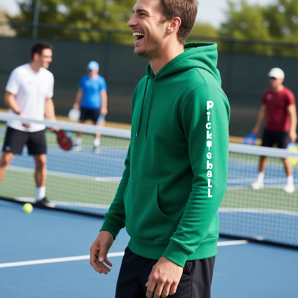 A man wearing a green long-sleeve unisex hoodie pullover with a white graphic text 'PICKLEBALL' on the sleeve with a pickleball paddle replacing the L, standing on a pickleball court.