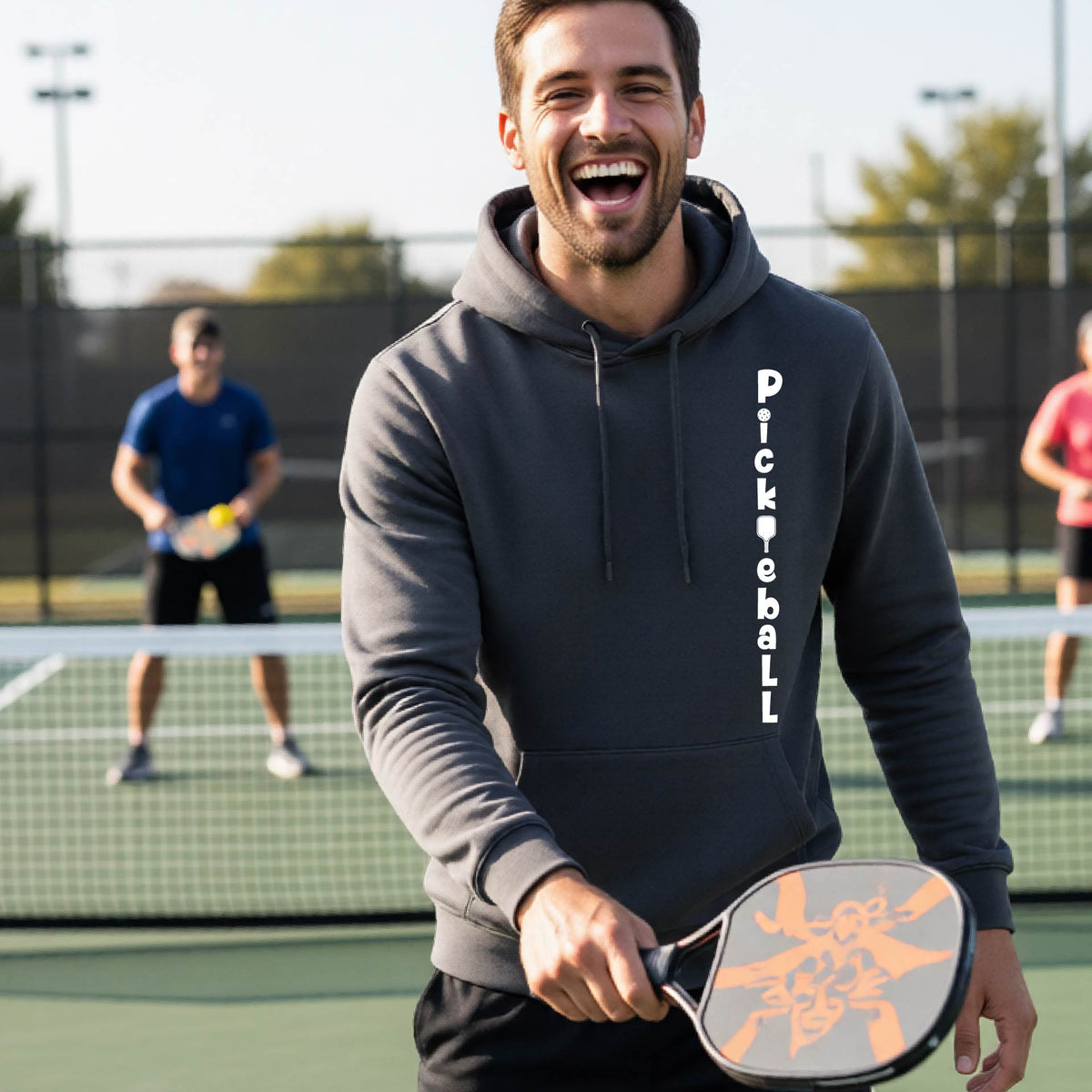 A man wearing a gray long-sleeve unisex hoodie pullover with a white graphic text 'PICKLEBALL' on the front with a pickleball paddle replacing the L, standing on a pickleball court.