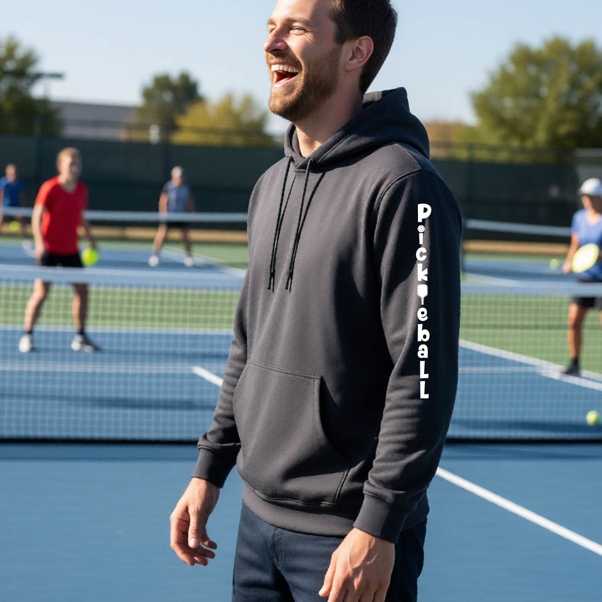 A man wearing a gray long-sleeve unisex hoodie pullover with a white graphic text 'PICKLEBALL' on the sleeve with a pickleball paddle replacing the L, standing on a pickleball court.