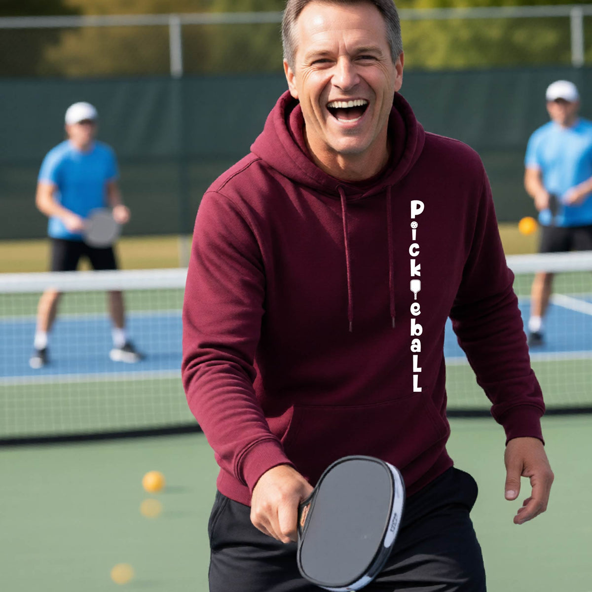 A man wearing a maroon long-sleeve unisex hoodie pullover with a white graphic text 'PICKLEBALL' on the front with a pickleball paddle replacing the L, standing on a pickleball court.