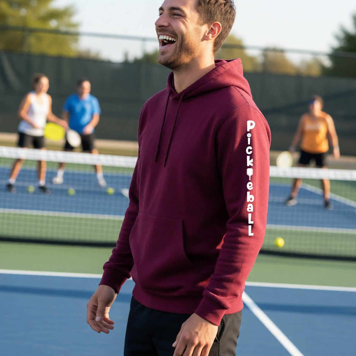 A man wearing a maroon long-sleeve unisex hoodie pullover with a white graphic text 'PICKLEBALL' on the sleeve with a pickleball paddle replacing the L, standing on a pickleball court.