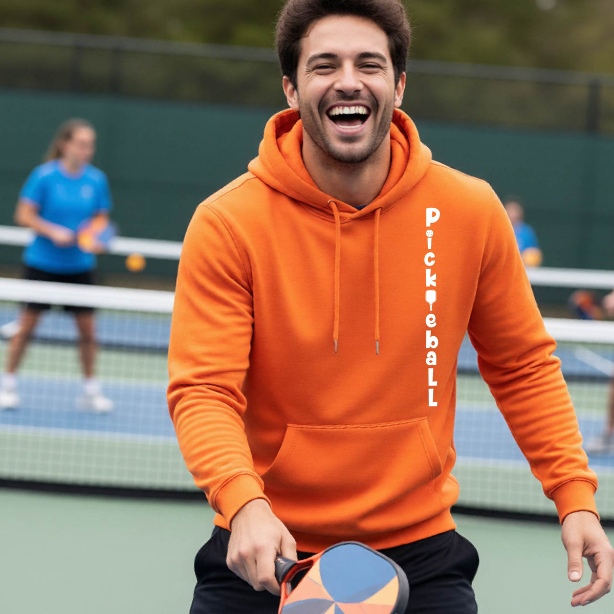 A man wearing an orange long-sleeve unisex hoodie pullover with a white graphic text 'PICKLEBALL' on the front with a pickleball paddle replacing the L, standing on a pickleball court.