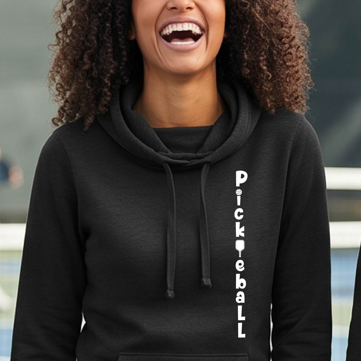 A woman wearing a black long-sleeve cowl-neck hoodie pullover with a white graphic text 'PICKLEBALL' on the front with a pickleball paddle replacing the L, standing on a pickleball court.