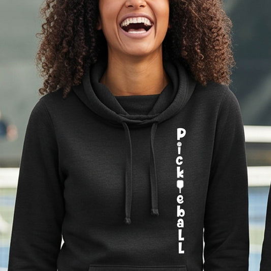 A woman wearing a black long-sleeve cowl-neck hoodie pullover with a white graphic text 'PICKLEBALL' on the front with a pickleball paddle replacing the L, standing on a pickleball court.