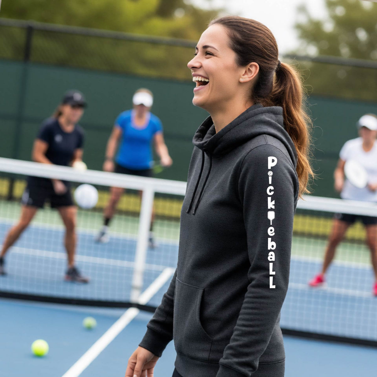 A woman wearing a black long-sleeve cowl-neck hoodie pullover with a white graphic text 'PICKLEBALL' on the sleeve with a pickleball paddle replacing the L, standing on a pickleball court.