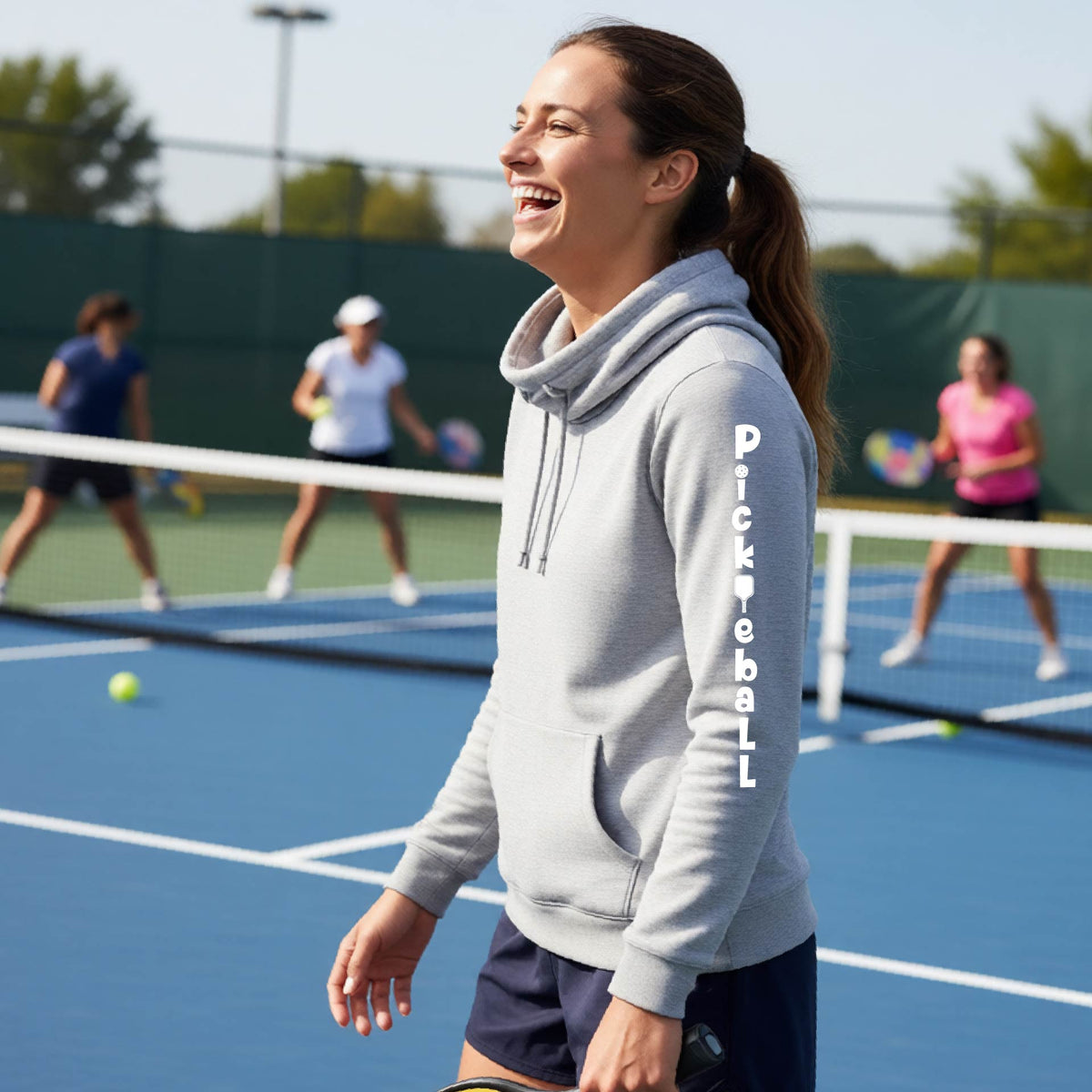 A woman wearing a gray long-sleeve cowl-neck hoodie pullover with a white graphic text 'PICKLEBALL' on the sleeve with a pickleball paddle replacing the L, standing on a pickleball court.