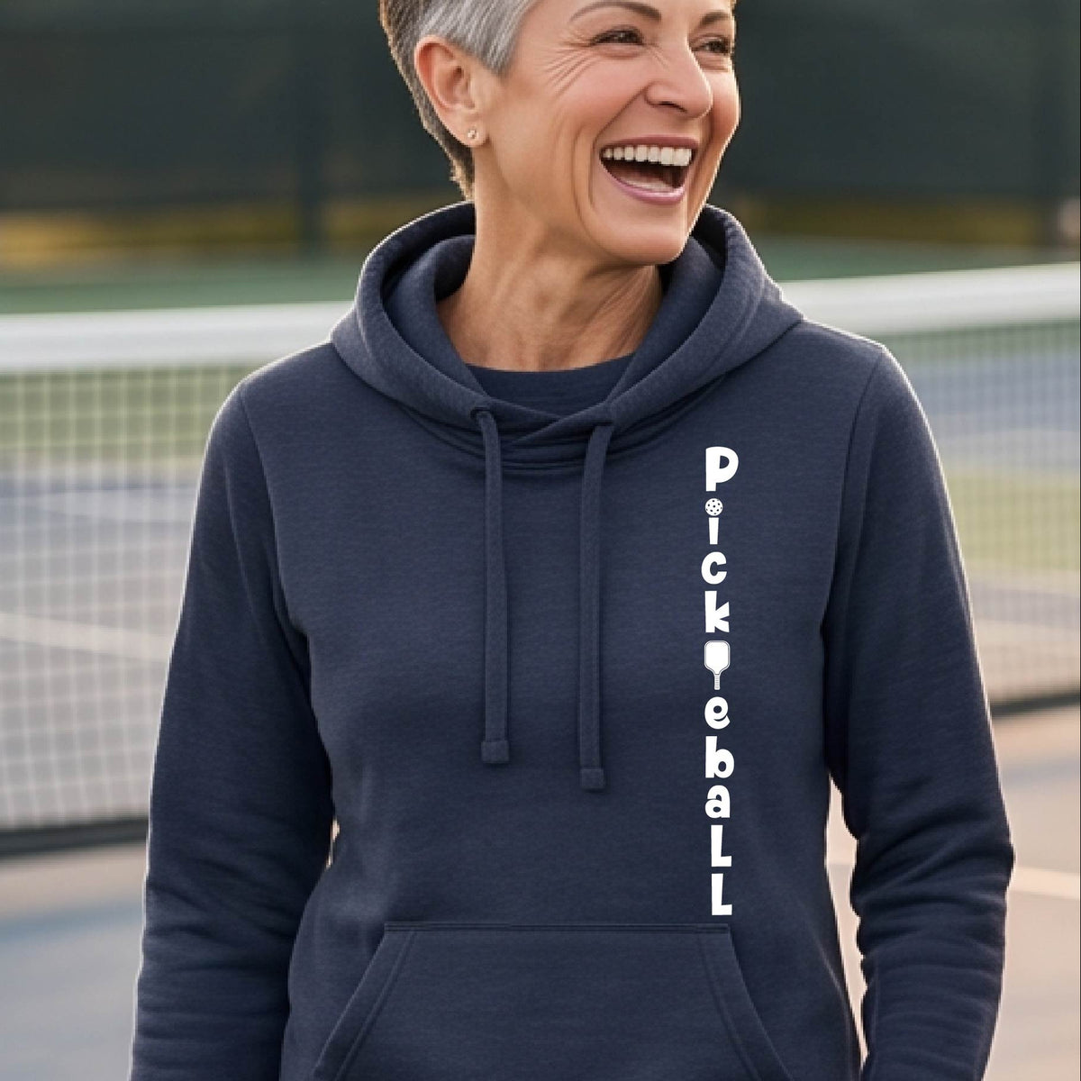 A woman wearing a navy blue long-sleeve cowl-neck hoodie pullover with a white graphic text 'PICKLEBALL' on the front with a pickleball paddle replacing the L, standing on a pickleball court.