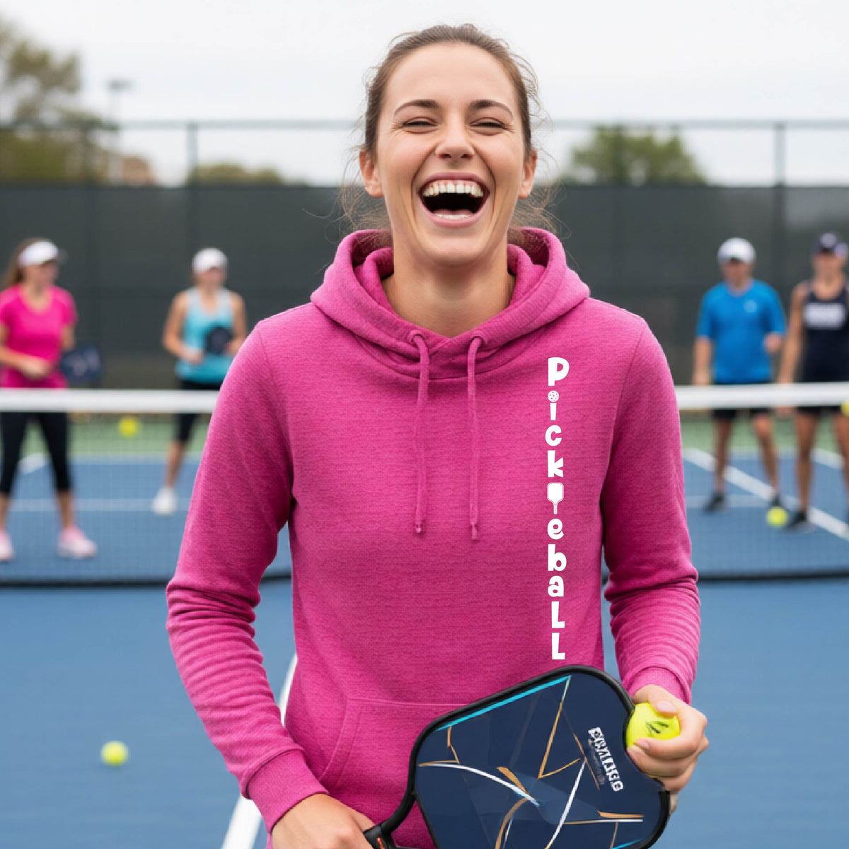 A woman wearing a pink long-sleeve cowl-neck hoodie pullover with a white graphic text 'PICKLEBALL' on the front with a pickleball paddle replacing the L, standing on a pickleball court.