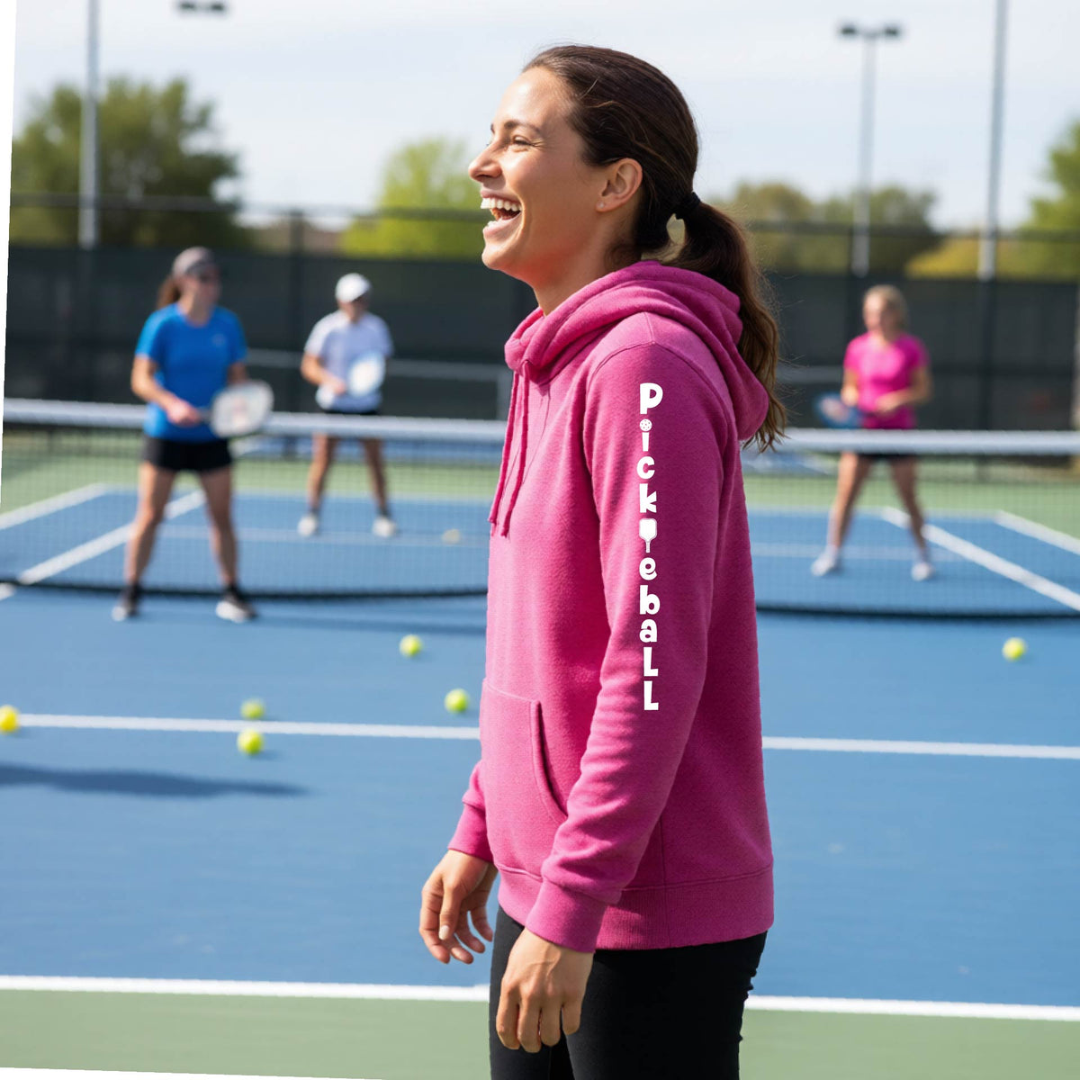 A woman wearing a pink long-sleeve cowl-neck hoodie pullover with a white graphic text 'PICKLEBALL' on the sleeve with a pickleball paddle replacing the L, standing on a pickleball court.