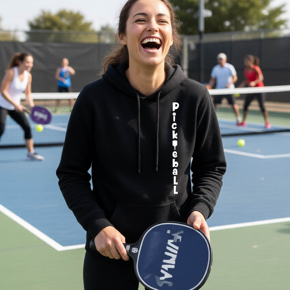 A woman wearing a black long-sleeve fitted hoodie pullover with a white graphic text 'PICKLEBALL' on the front with a pickleball paddle replacing the L, standing on a pickleball court.