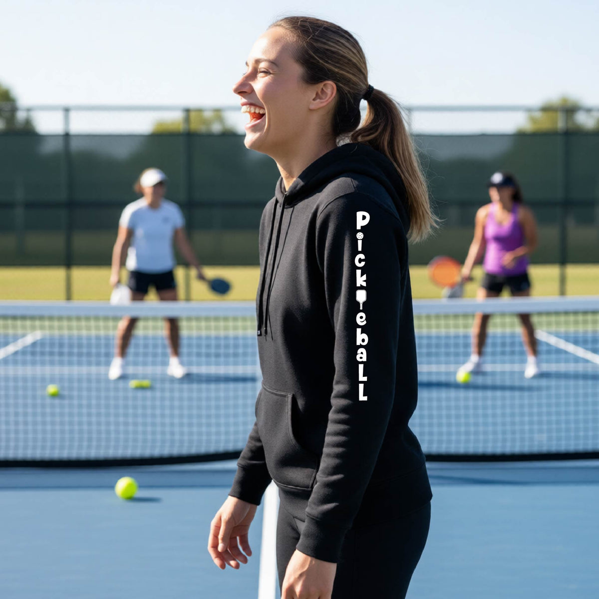 A woman wearing a black long-sleeve fitted hoodie pullover with a white graphic text 'PICKLEBALL' on the sleeve with a pickleball paddle replacing the L, standing on a pickleball court.
