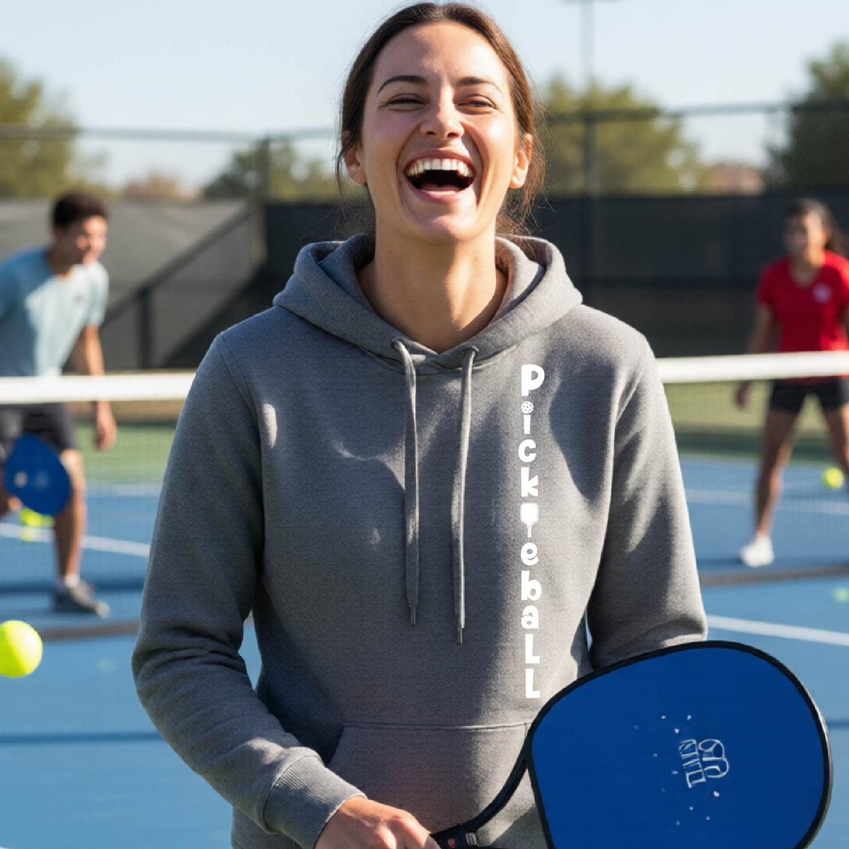 A woman wearing a gray long-sleeve fitted hoodie pullover with a white graphic text 'PICKLEBALL' on the front with a pickleball paddle replacing the L, standing on a pickleball court.