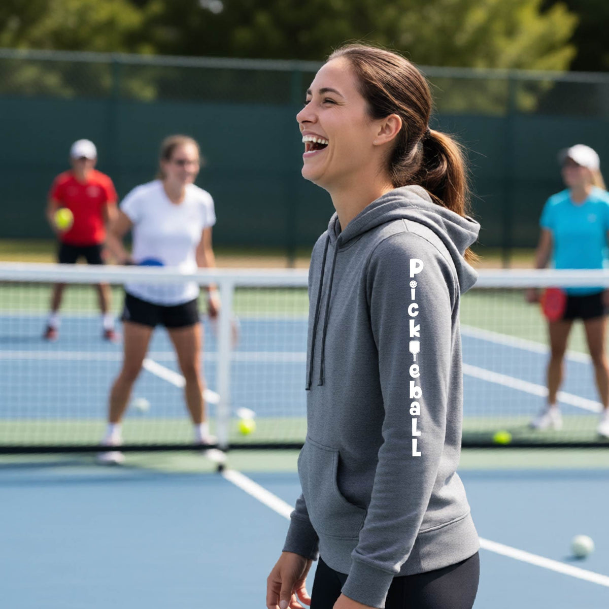 A woman wearing a gray long-sleeve fitted hoodie pullover with a white graphic text 'PICKLEBALL' on the sleeve with a pickleball paddle replacing the L, standing on a pickleball court.