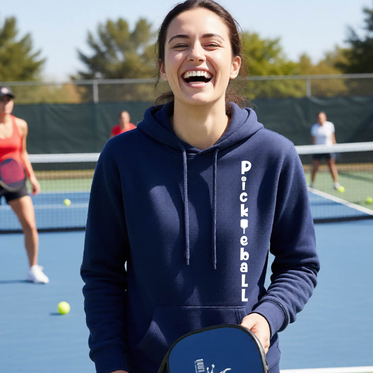 A woman wearing a navy blue long-sleeve fitted hoodie pullover with a white graphic text 'PICKLEBALL' on the front with a pickleball paddle replacing the L, standing on a pickleball court.