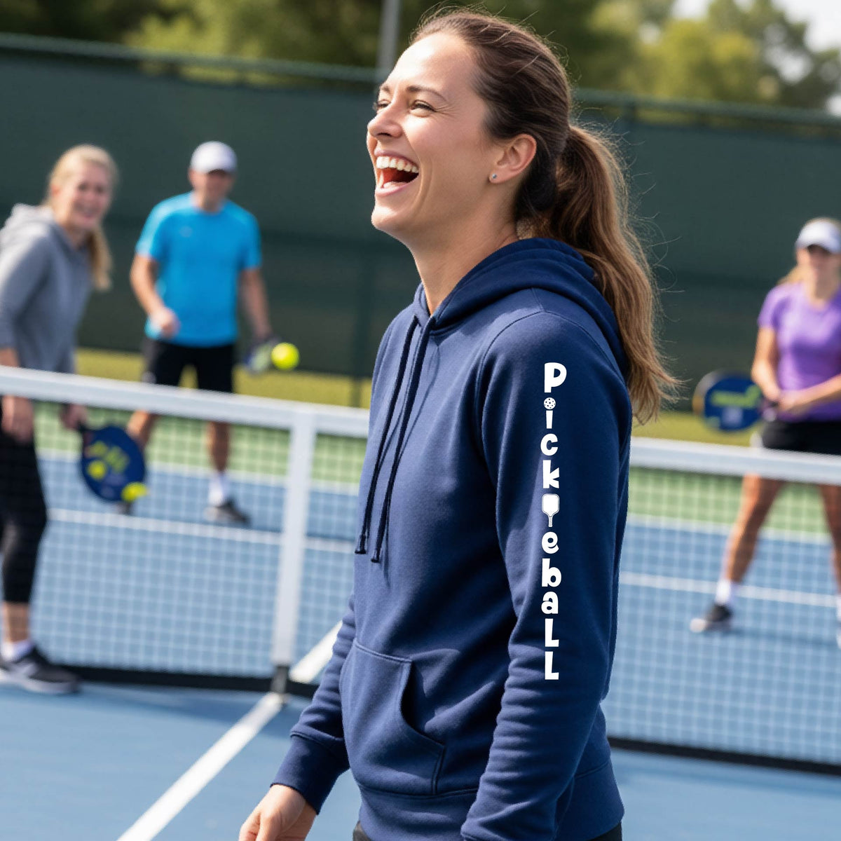  A woman wearing a navy blue long-sleeve fitted hoodie pullover with a white graphic text 'PICKLEBALL' on the sleeve with a pickleball paddle replacing the L, standing on a pickleball court.