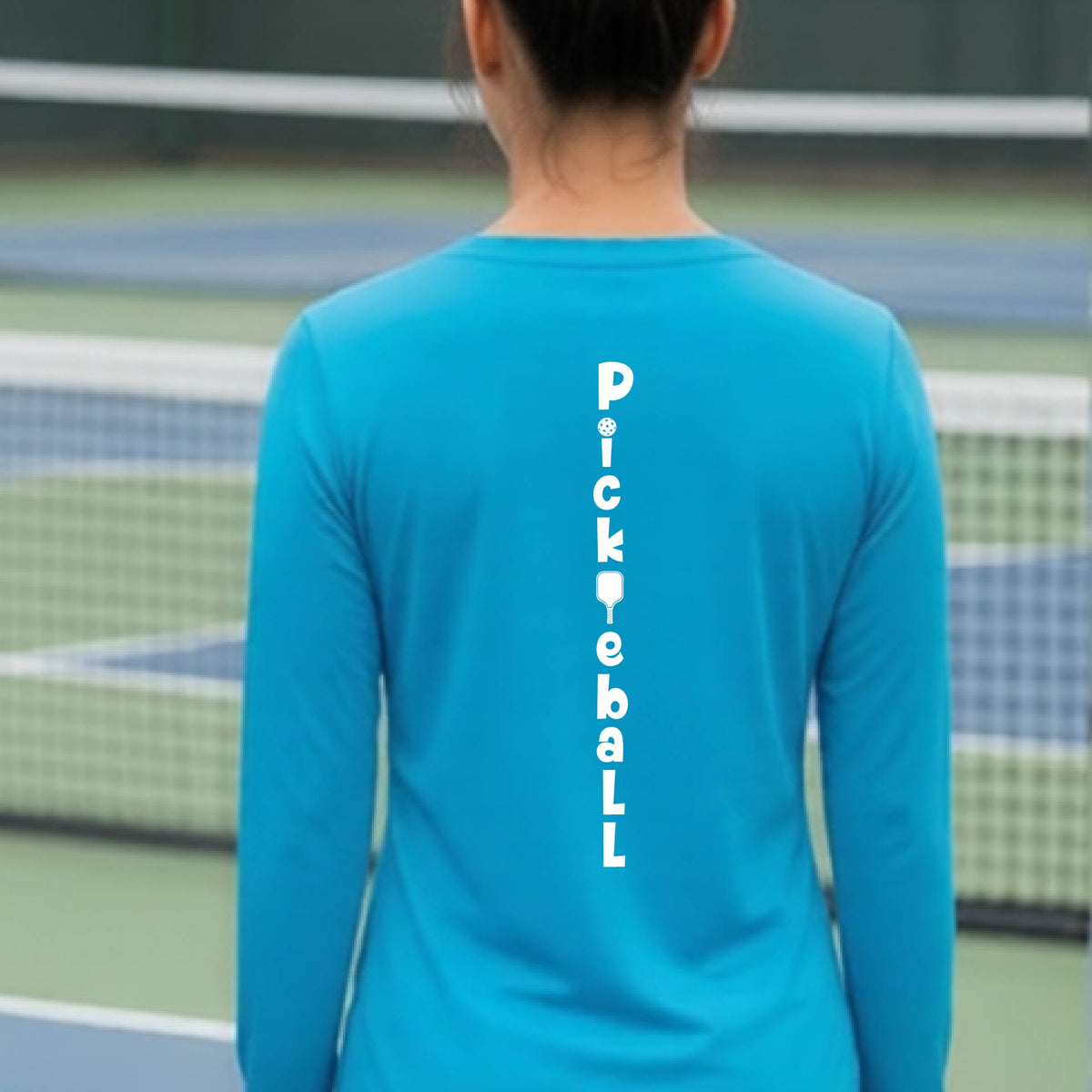 A woman wearing a bright blue long-sleeve t-shirt with a white graphic text 'PICKLEBALL' on the back with a pickleball paddle replacing the L, standing on a pickleball court.