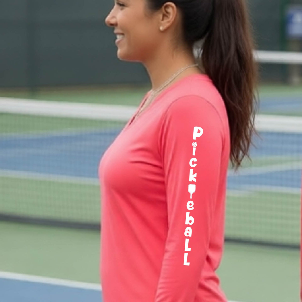 A woman wearing a coral pink long-sleeve t-shirt with a white graphic text 'PICKLEBALL' on the sleeve with a pickleball paddle replacing the L, standing on a pickleball court.
