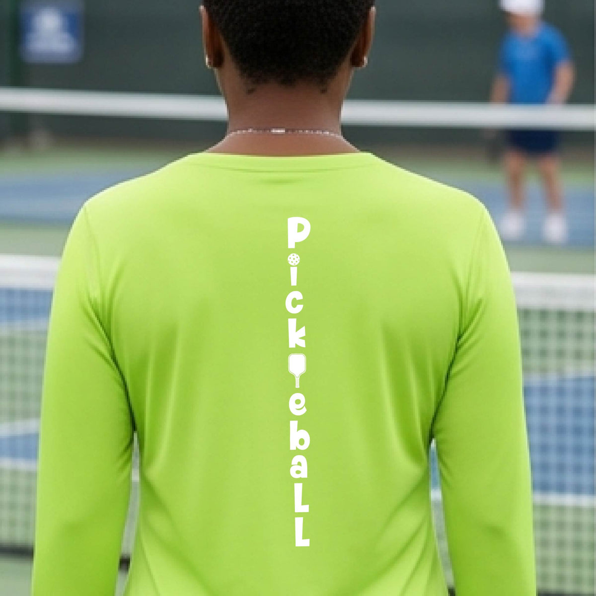 A woman wearing a lime green long-sleeve t-shirt with a white graphic text 'PICKLEBALL' on the back with a pickleball paddle replacing the L, standing on a pickleball court.