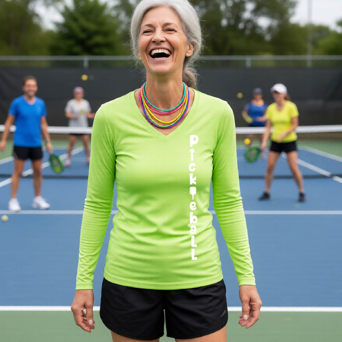 A woman wearing a lime green long-sleeve t-shirt with a white graphic text 'PICKLEBALL' on the front with a pickleball paddle replacing the L, standing on a pickleball court.