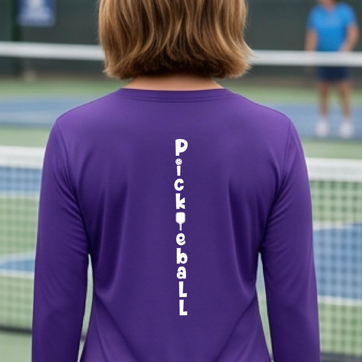 A woman wearing a purple long-sleeve t-shirt with a white graphic text 'PICKLEBALL' on the back with a pickleball paddle replacing the L, standing on a pickleball court.