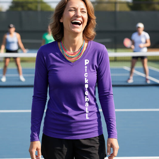 A woman wearing a purple long-sleeve t-shirt with a white graphic text 'PICKLEBALL' on the front with a pickleball paddle replacing the L, standing on a pickleball court.