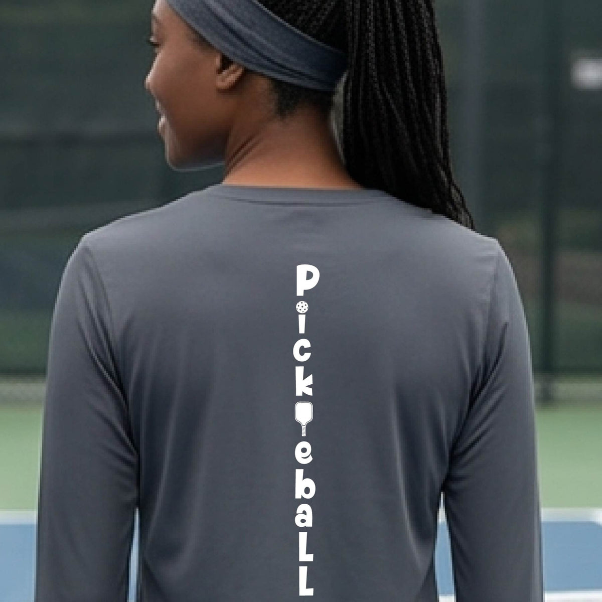 A woman wearing a gray long-sleeve t-shirt with a white graphic text 'PICKLEBALL' on the back with a pickleball paddle replacing the L, standing on a pickleball court.