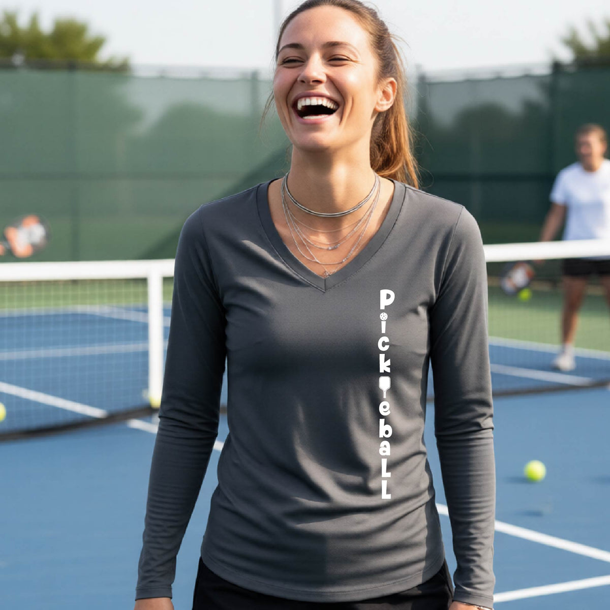 A woman wearing a gray long-sleeve t-shirt with a white graphic text 'PICKLEBALL' on the front with a pickleball paddle replacing the L, standing on a pickleball court.