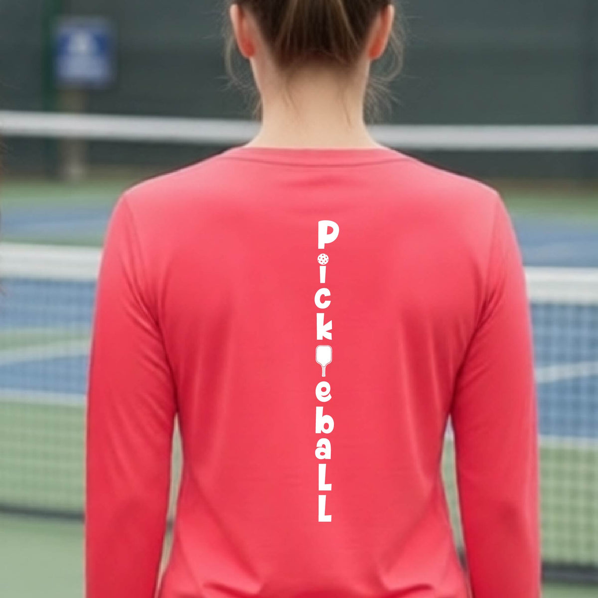 A woman wearing a coral pink long-sleeve t-shirt with a white graphic text 'PICKLEBALL' on the back with a pickleball paddle replacing the L, standing on a pickleball court.