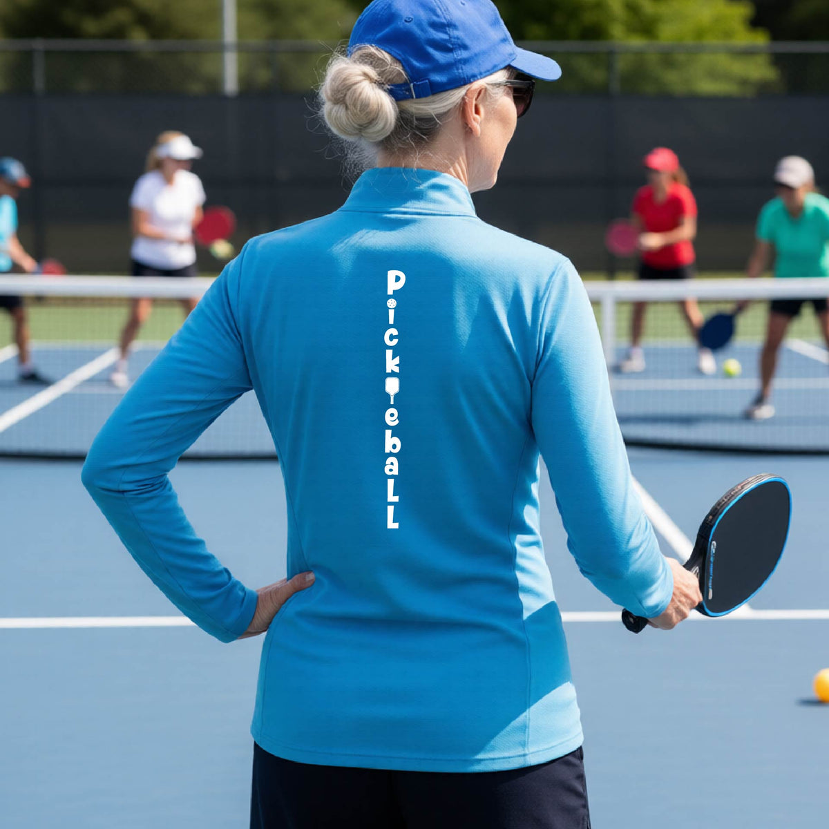 A woman wearing a bright blue quarter-zip long-sleeve t-shirt with a white graphic text 'PICKLEBALL' on the back with a pickleball paddle replacing the L, standing on a pickleball court. 