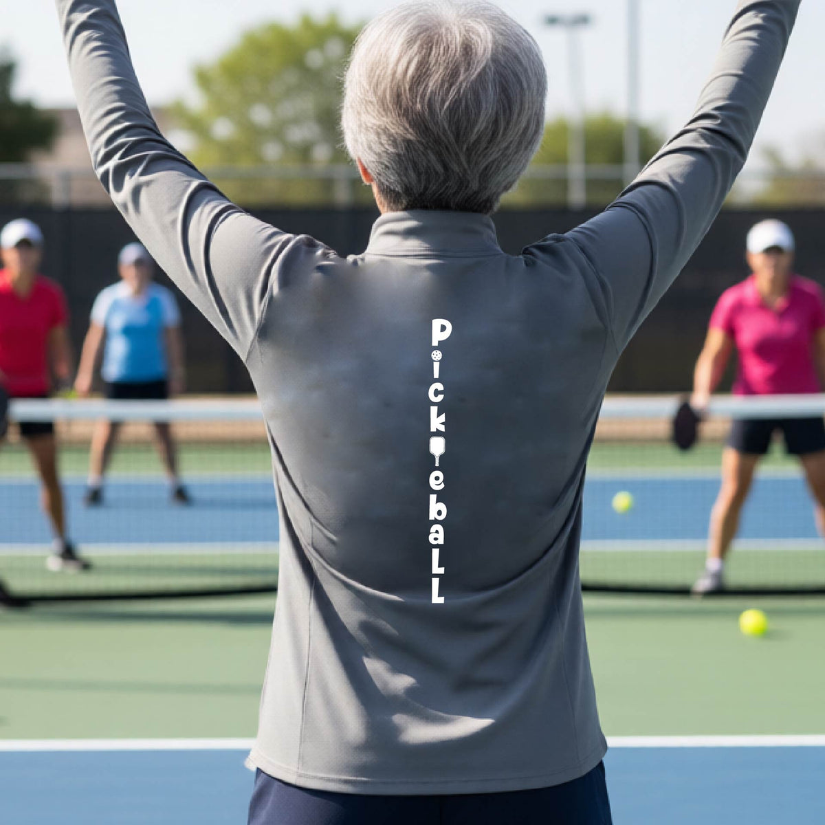 A woman wearing a gray quarter-zip long-sleeve t-shirt with a white graphic text 'PICKLEBALL' on the back with a pickleball paddle replacing the L, standing on a pickleball court.