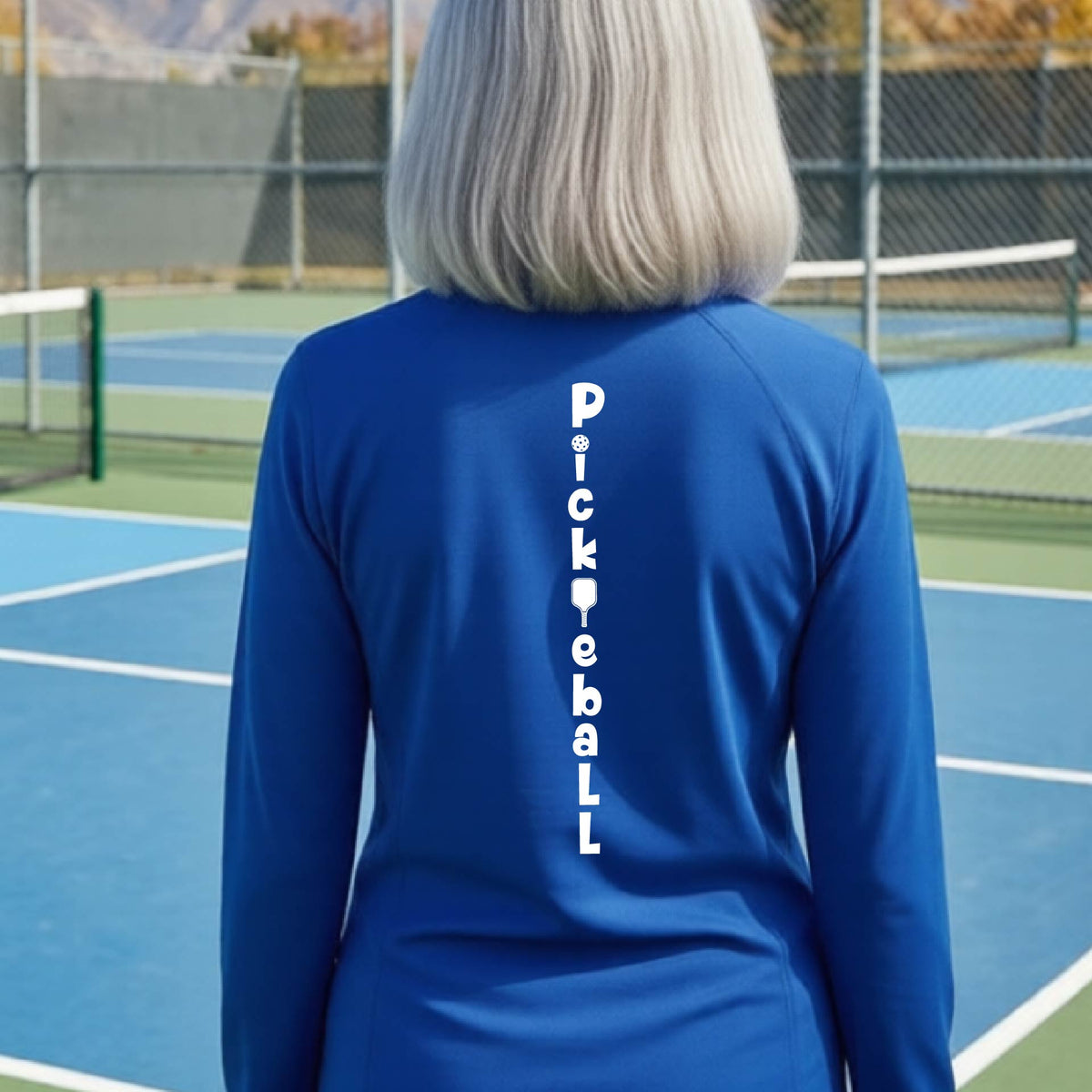 A woman wearing a royal blue quarter-zip long-sleeve t-shirt with a white graphic text 'PICKLEBALL' on the back with a pickleball paddle replacing the L, standing on a pickleball court.