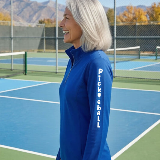 A woman wearing a royal blue quarter-zip long-sleeve t-shirt with a white graphic text 'PICKLEBALL' on the sleeve with a pickleball paddle replacing the L, standing on a pickleball court.
