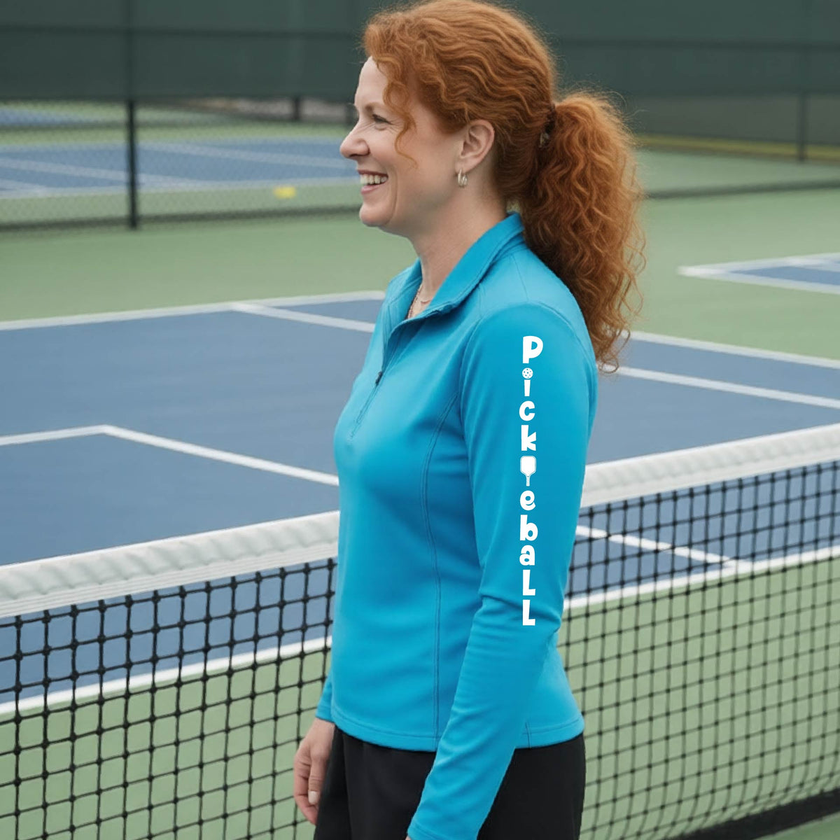 A woman wearing a bright blue quarter-zip long-sleeve t-shirt with a white graphic text 'PICKLEBALL' on the sleeve with a pickleball paddle replacing the L, standing on a pickleball court.