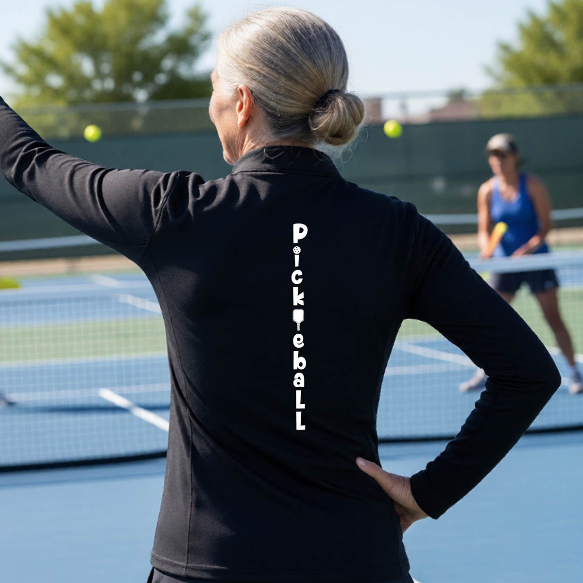 A woman wearing a black quarter-zip long-sleeve t-shirt with a white graphic text 'PICKLEBALL' on the back with a pickleball paddle replacing the L, standing on a pickleball court.