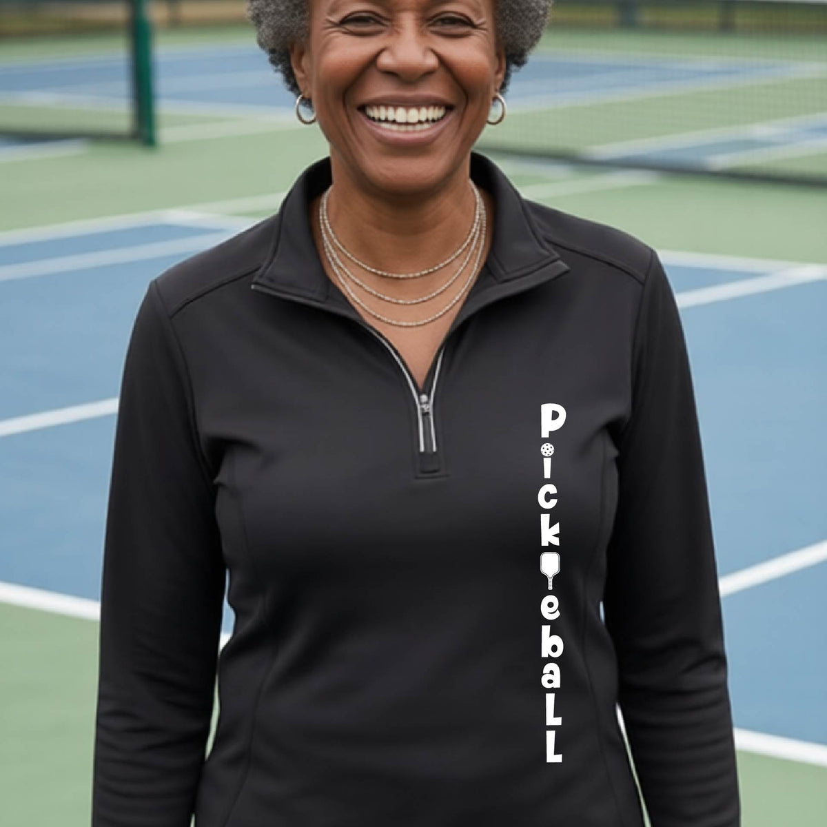 A woman wearing a black quarter-zip long-sleeve t-shirt with a white graphic text 'PICKLEBALL' on the front with a pickleball paddle replacing the L, standing on a pickleball court.